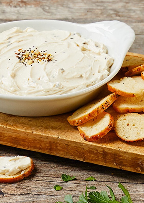Close-up of a creamy white dip in a shallow serving dish, topped with seasoning, set on a wooden board with sliced baguette rounds, one piece spread with the dip in the foreground.