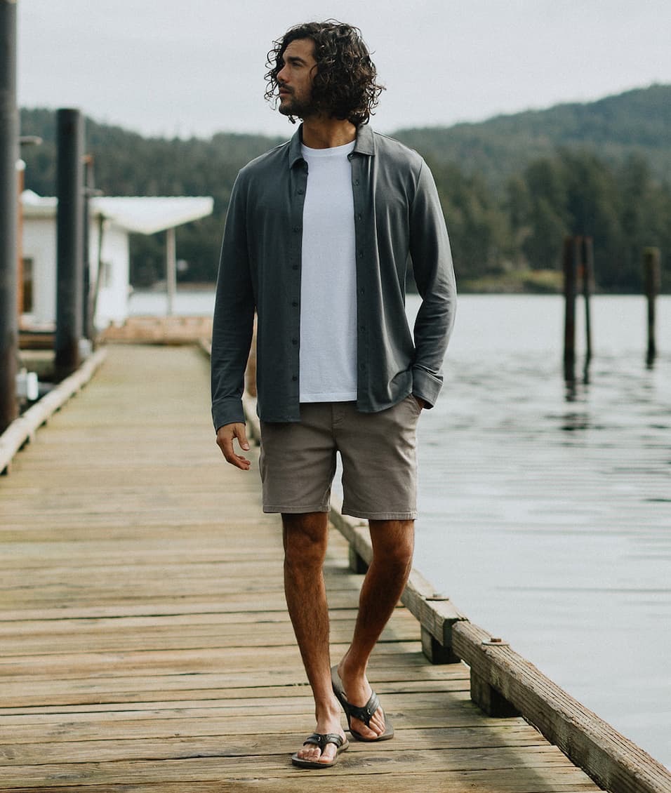 Man walking along a wooden dock in a relaxed outfit—lightweight button-down over a white tee, shorts, and sandals—with a calm lakeside setting in the background.