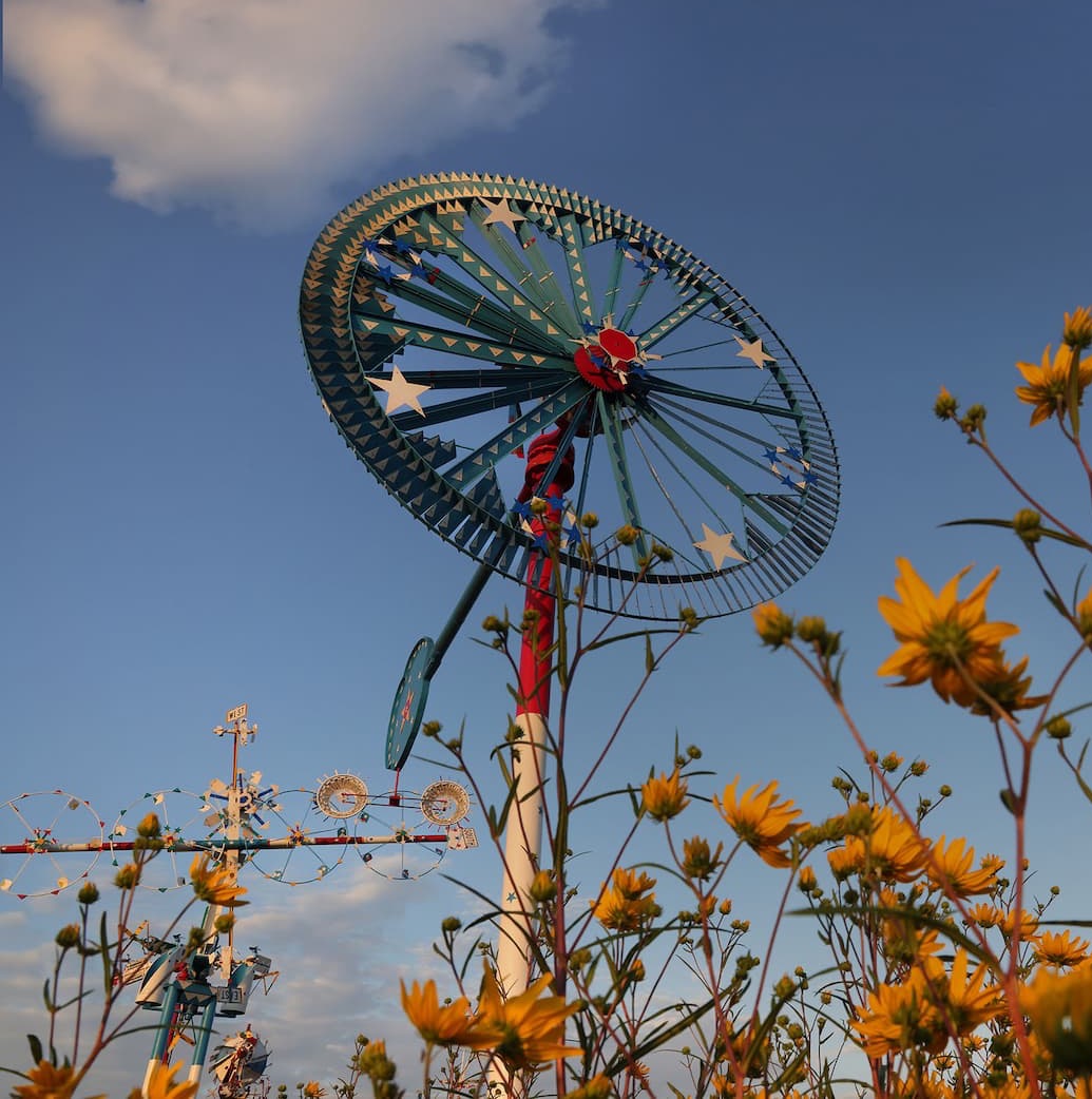 Colorful whirligig wind sculptures rising above yellow flowers against a blue sky.