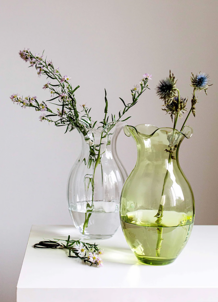 Two glass vases, one clear and one soft green, hold delicate wildflowers on a simple tabletop, creating a light, minimalist arrangement.
