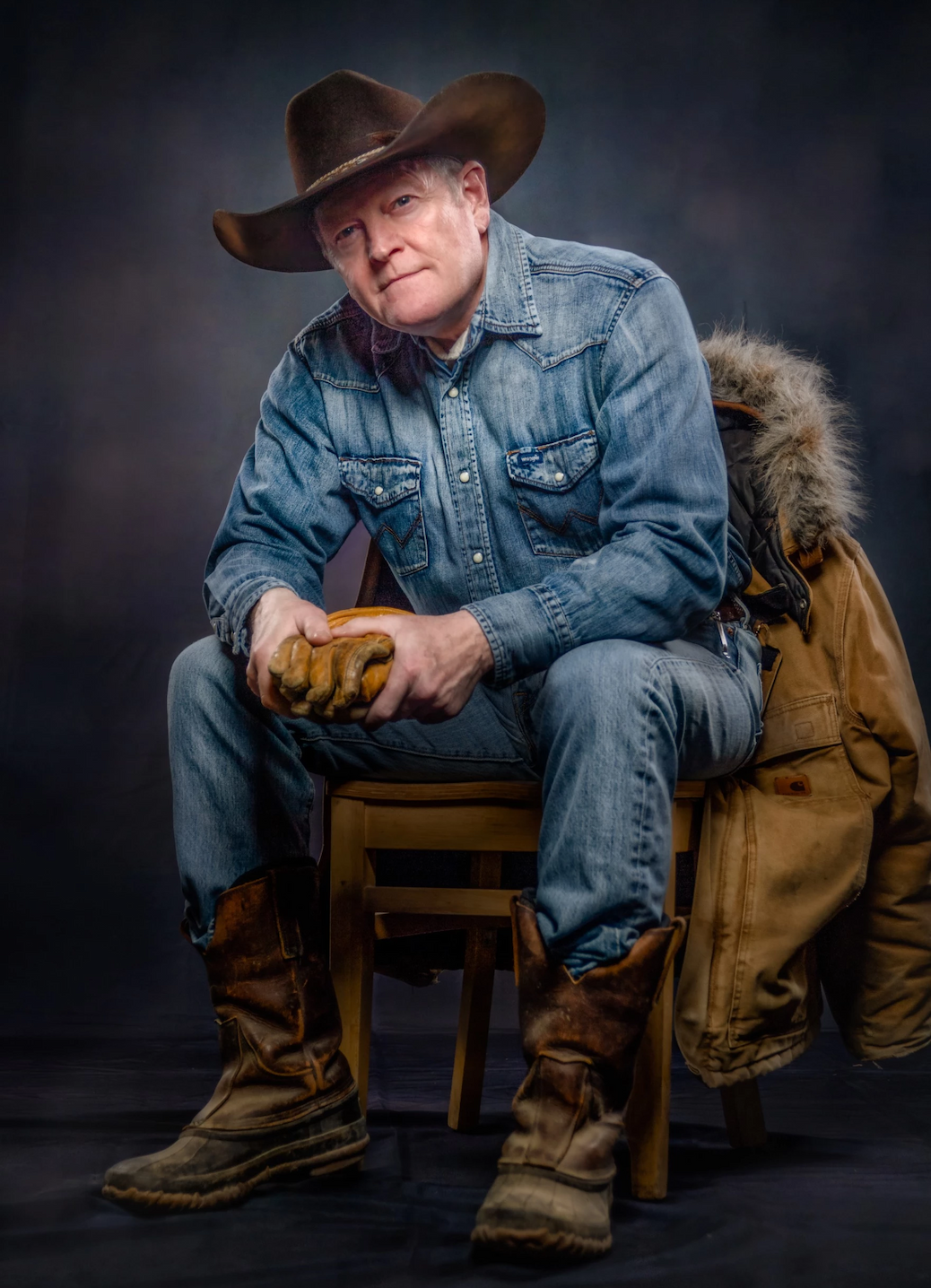 Portrait of a man seated on a wooden chair wearing a denim western shirt, jeans, rugged boots, and a wide-brim cowboy hat. Work gloves rest in his hands, and a heavy canvas coat with a fur-lined hood hangs over the chair, creating a rugged ranch-inspired look against a dark studio backdrop.