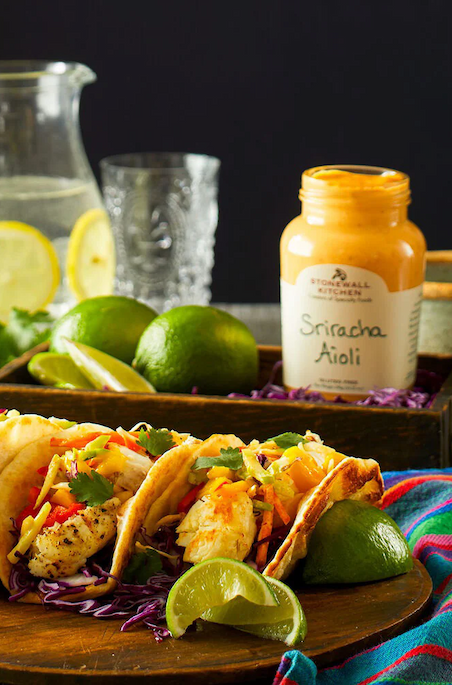 Three fish tacos topped with shredded cabbage, cilantro, and cheese, served with lime wedges and a jar of Stonewall Kitchen Sriracha Aioli, with limes and a pitcher of lemon water in the background.