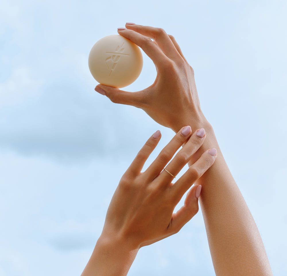 Two hands with neatly manicured nails are held up against a blue sky, one hand gracefully holding a round, beige object with an embossed logo. The other hand is gently touching the wrist. Fearrington Village