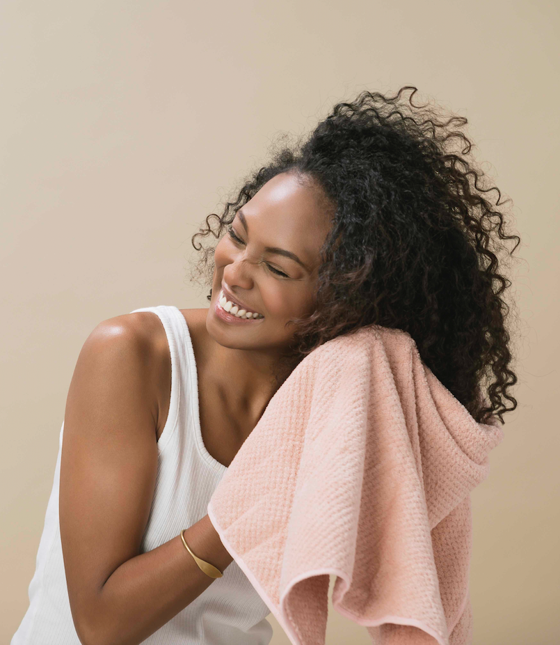 Woman with natural curly hair smiling while drying it with a soft pink towel, wearing a white tank top against a neutral beige background.