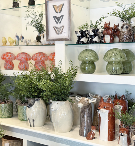 Shelving display in a boutique with whimsical ceramic vases and figurines shaped like animals and mushrooms, soft green plants tucked among them, and a framed butterfly print above, creating a playful, nature-inspired arrangement.