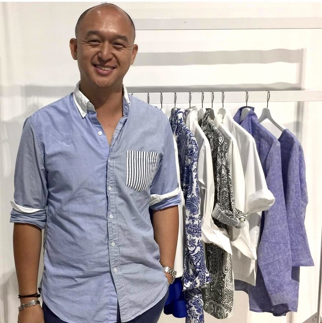 A smiling man stands beside a clothing rack displaying neatly hung shirts and patterned garments in shades of blue.