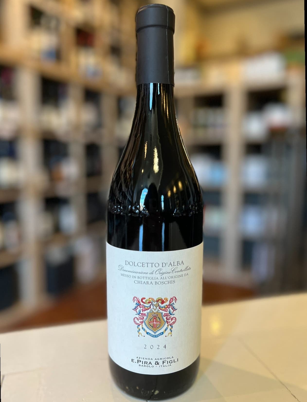 Bottle of E. Pira & Figli Dolcetto d’Alba 2024 wine standing on a counter, with softly blurred shelves of bottles behind it.