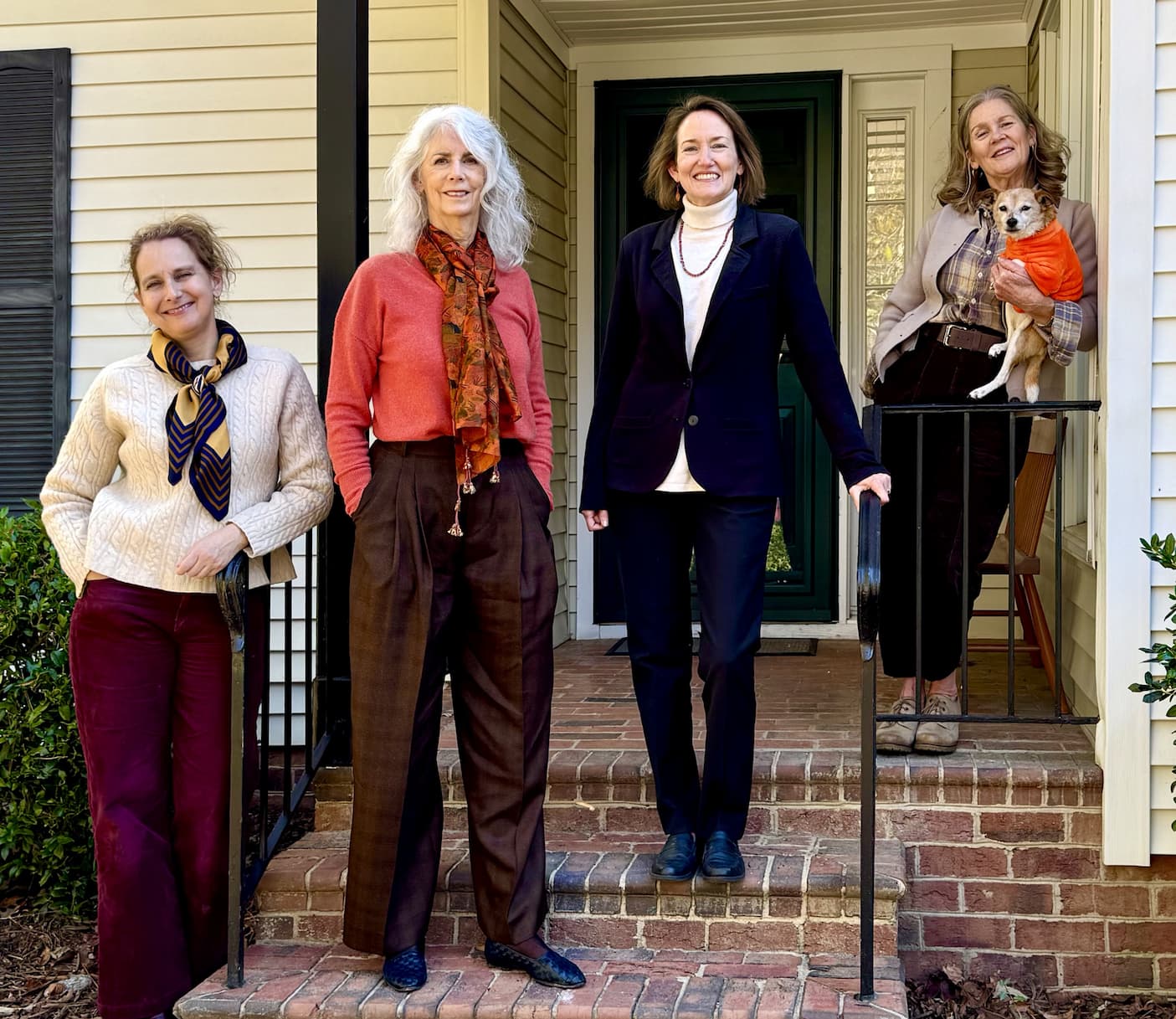 Four women pose together on the steps of a brick porch in front of a house; one woman holds a small dog wearing an orange sweater. All are smiling and dressed in casual, stylish clothes. Fearrington Village