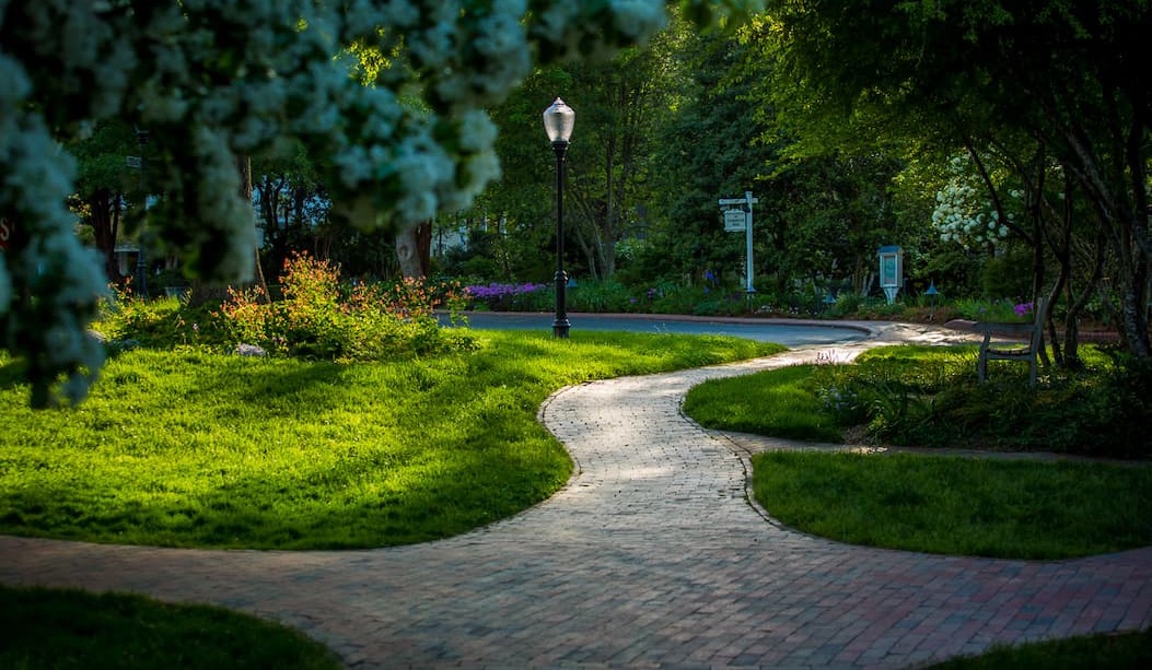 Curving brick pathway winding through lush green lawns, flowering trees, and landscaped gardens in soft morning light. Fearrington Village.