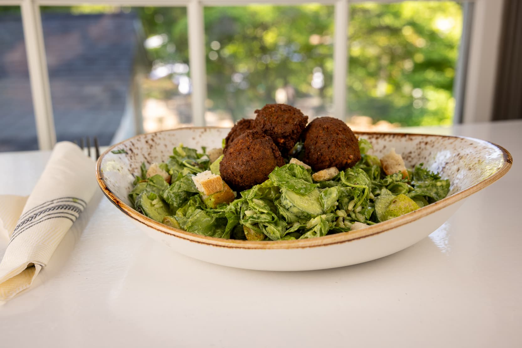 A rustic ceramic bowl filled with crisp greens sits on a bright table near a sunlit window. The salad is topped with several golden-brown falafel, avocado slices, croutons, and grains, all lightly dressed and arranged for a fresh, colorful presentation. A folded linen napkin and fork rest beside the bowl, while the soft-focus garden view beyond the window creates a relaxed, inviting dining atmosphere.