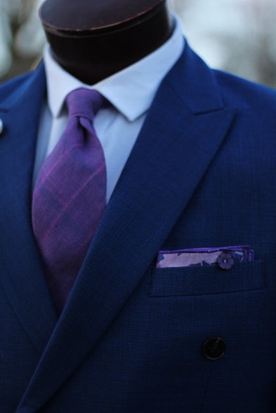 Close-up of a tailored blue double-breasted suit on a mannequin, paired with a white shirt, purple tie, and matching pocket square.