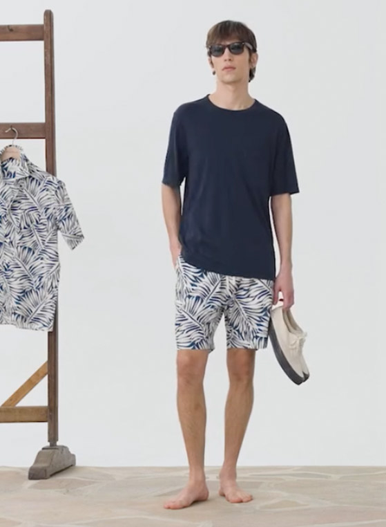 Man wearing a navy T-shirt with tropical print shorts, holding slip-on shoes and sunglasses. Fearrington Village.