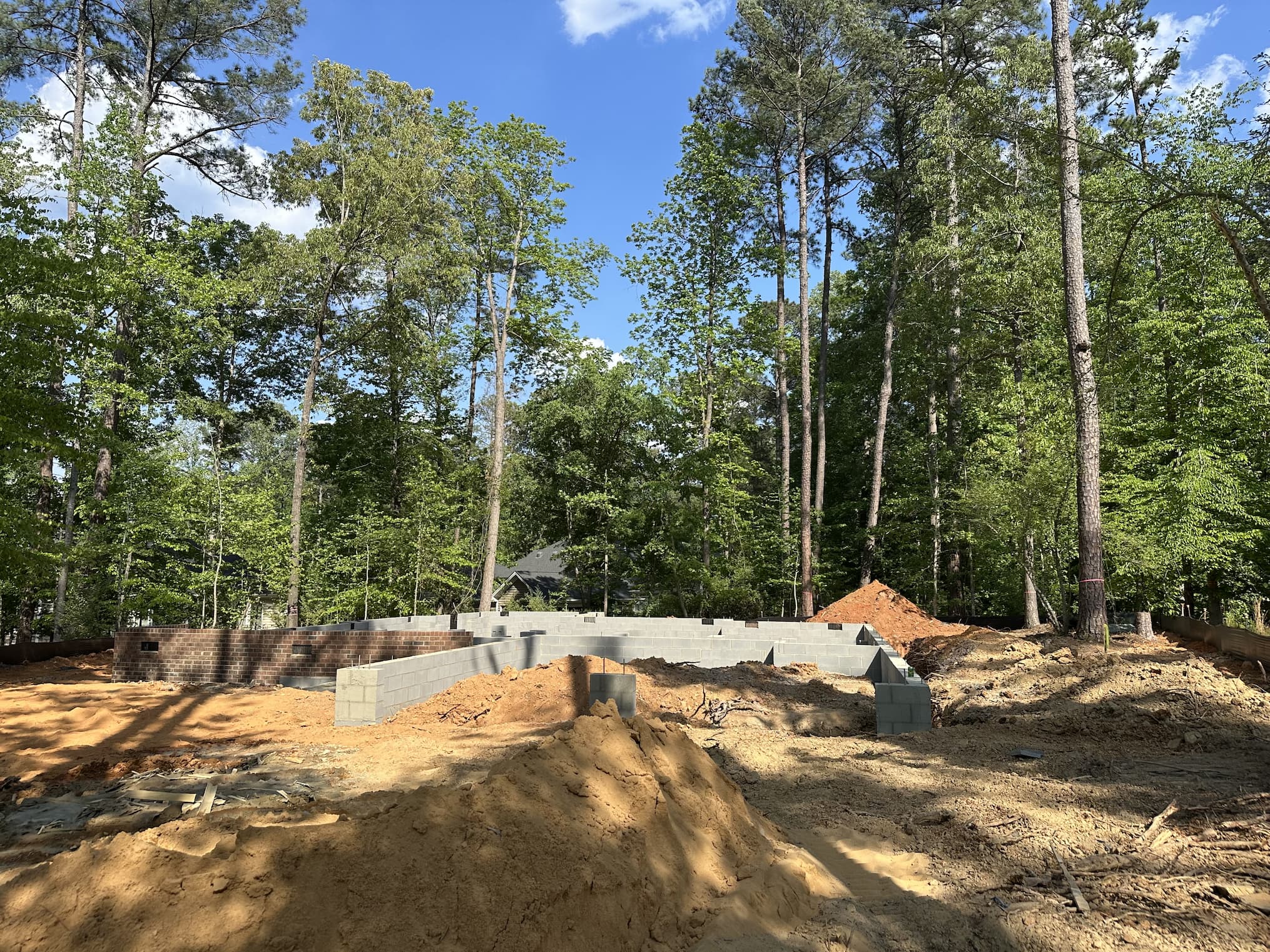 Construction site in a wooded area with a newly laid concrete block foundation, piles of dirt, and tall trees surrounding the future home under a clear blue sky.