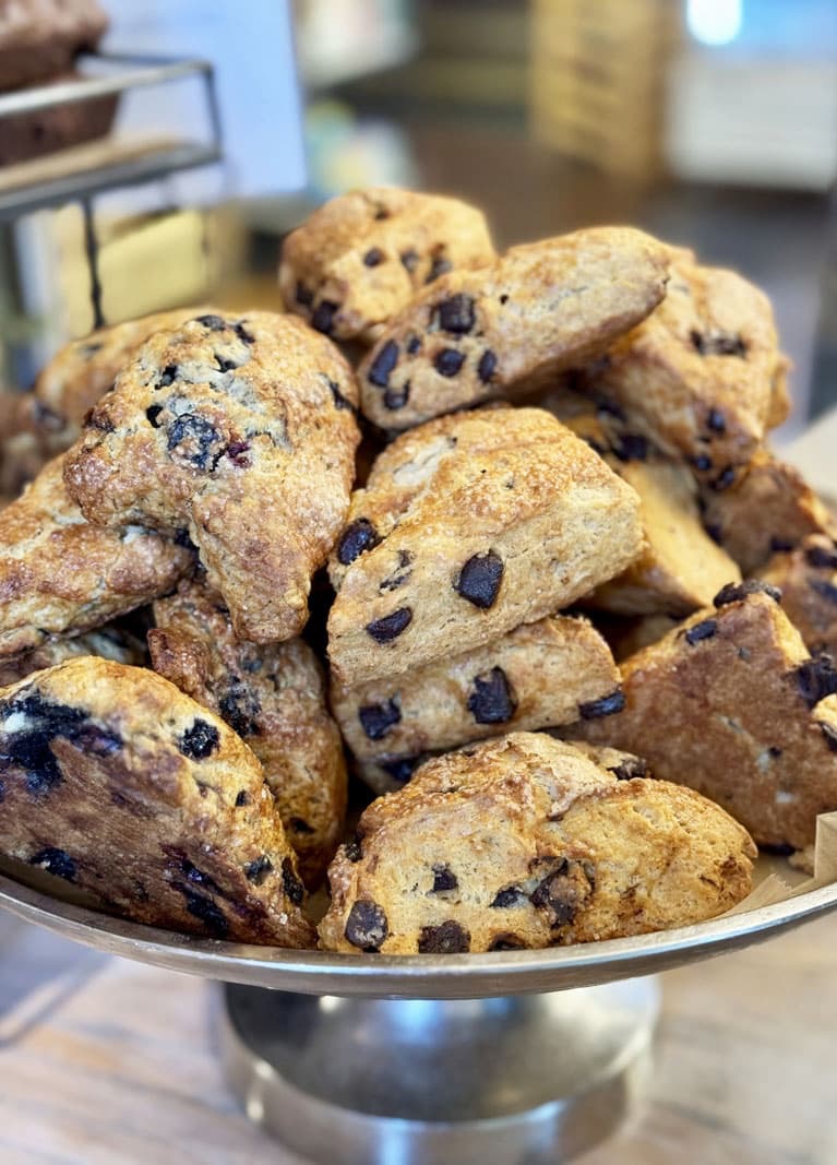 Pile of golden-brown chocolate chip scones stacked on a metal stand, with crisp edges and visible chocolate chunks, set in a bakery display.