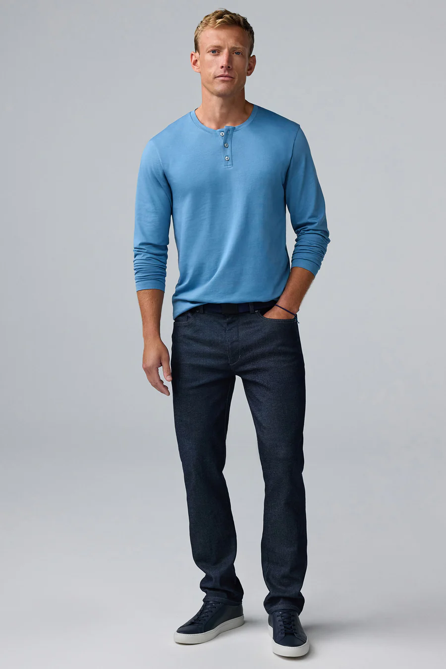 A man stands against a plain light background wearing a blue long-sleeve henley shirt, dark jeans, and sneakers, with one hand in his pocket.