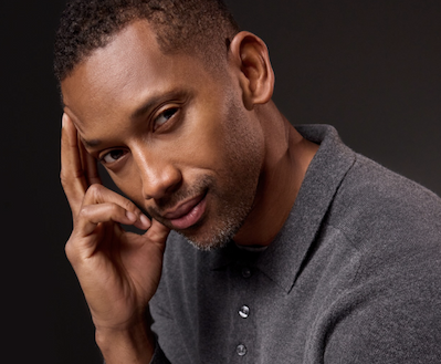 Close-up portrait of a man in a charcoal polo shirt, resting his head against his hand and looking directly at the camera with a calm, thoughtful expression against a dark, softly lit background.