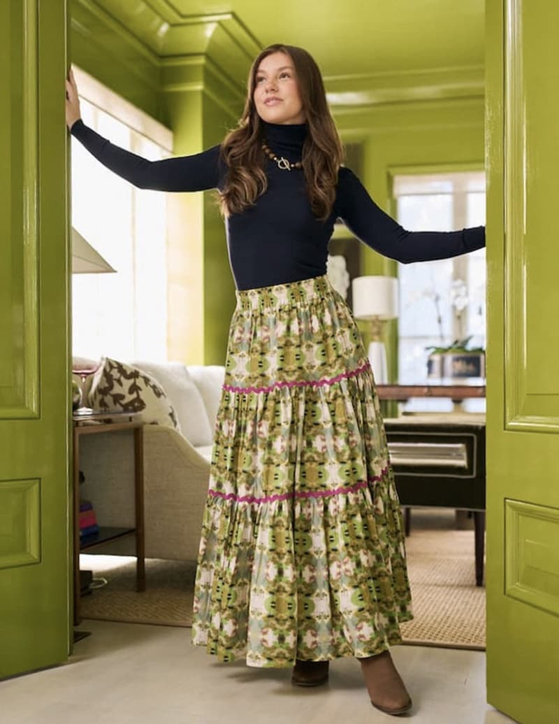 A woman stands in a doorway of a green room, wearing a long floral skirt, black turtleneck, and brown shoes. She gazes ahead with a relaxed pose, with a desk, lamp, and couches visible in the background. Fearrington Village