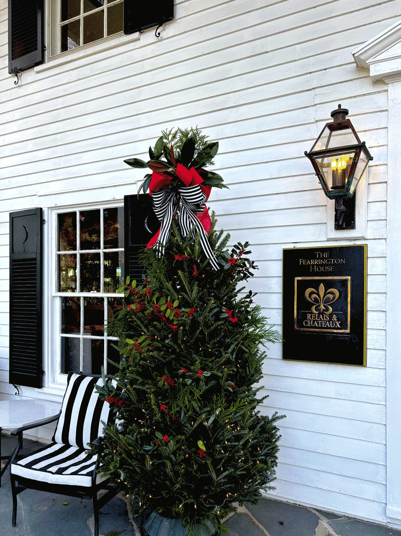 Decorated evergreen tree with lights, red accents, and a black-and-white ribbon bow, displayed outside The Fearrington House entrance beside a striped chair and a Relais & Châteaux sign on a white clapboard wall.
