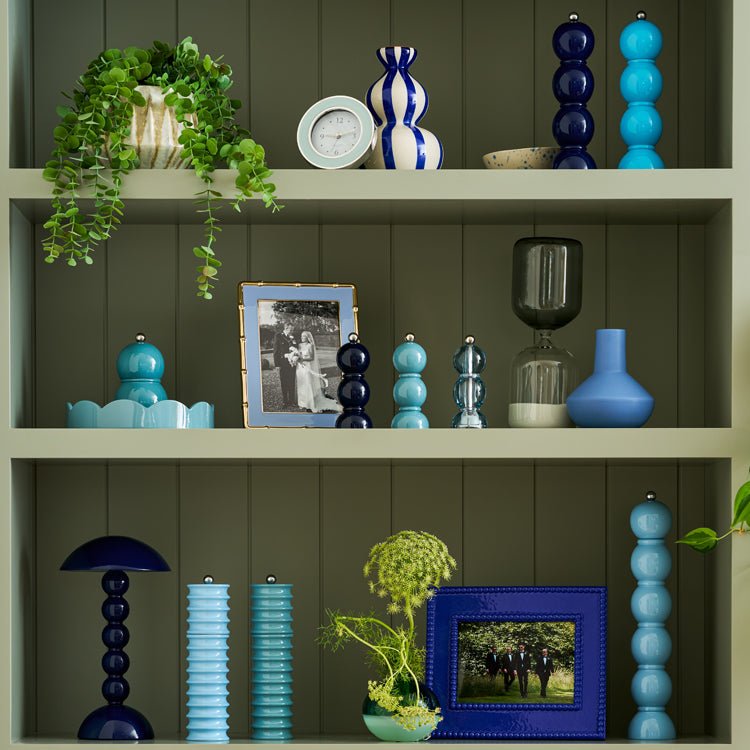 Green built-in shelving styled with layered blue and teal accents, including sculptural pepper mills, vases, and glass vessels, mixed with framed photos, a small clock, and trailing greenery for a balanced, modern display.