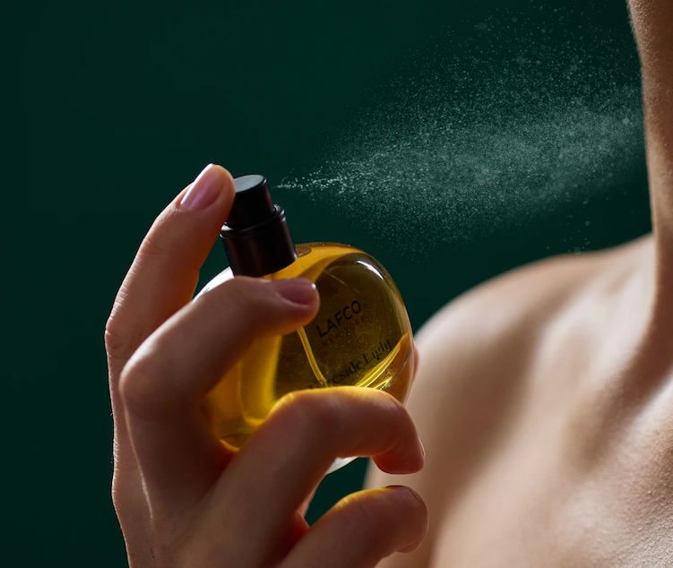 Close-up of LAFCO Fireside Light eau de parfum being sprayed onto skin, with a fine mist visible against a deep green background. Fearrington Village.