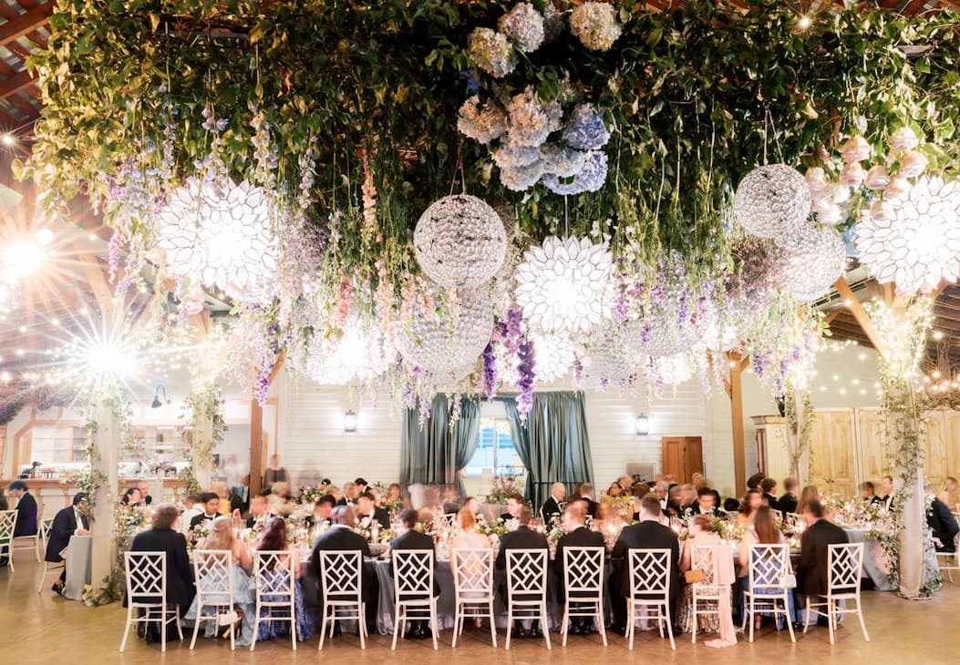 Wedding reception in a barn with long tables of guests beneath a dramatic ceiling installation of greenery, hanging flowers, and glowing white lanterns.