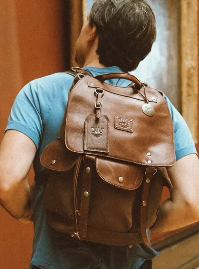Man seen from behind wearing a blue shirt and carrying a brown leather backpack with multiple pockets, straps, and metal hardware. The setting appears indoors with warm-toned walls, giving a rustic, travel-ready feel.