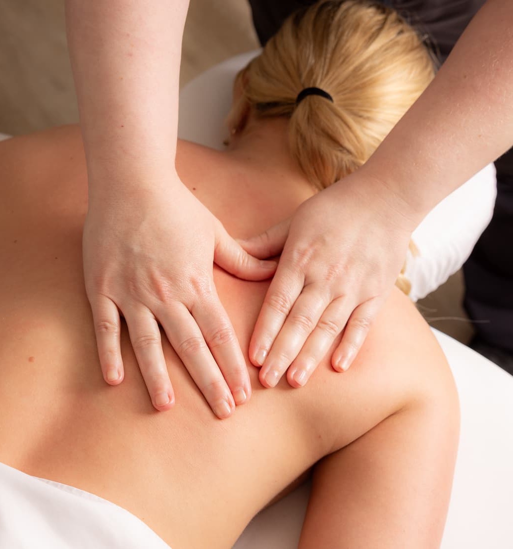 A massage therapist applies a relaxing back massage to a client lying face down on a treatment table. The therapist's hands press gently into the upper back and shoulder area, while the client rests comfortably with blonde hair tied back. The softly lit spa setting creates a calm, restorative atmosphere focused on relaxation and wellness.