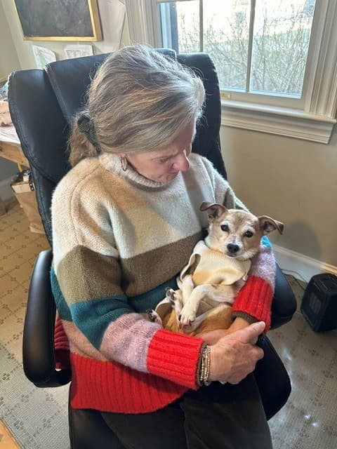 A woman seated in a home office cradles a small tan-and-white dog in her lap. She wears a color-block sweater in cream, green, pink, and red tones and looks down affectionately at the dog, which is wrapped in a light-colored blanket and gazes toward the camera. Natural light streams through a nearby window, creating a warm, intimate scene that conveys comfort, companionship, and care.