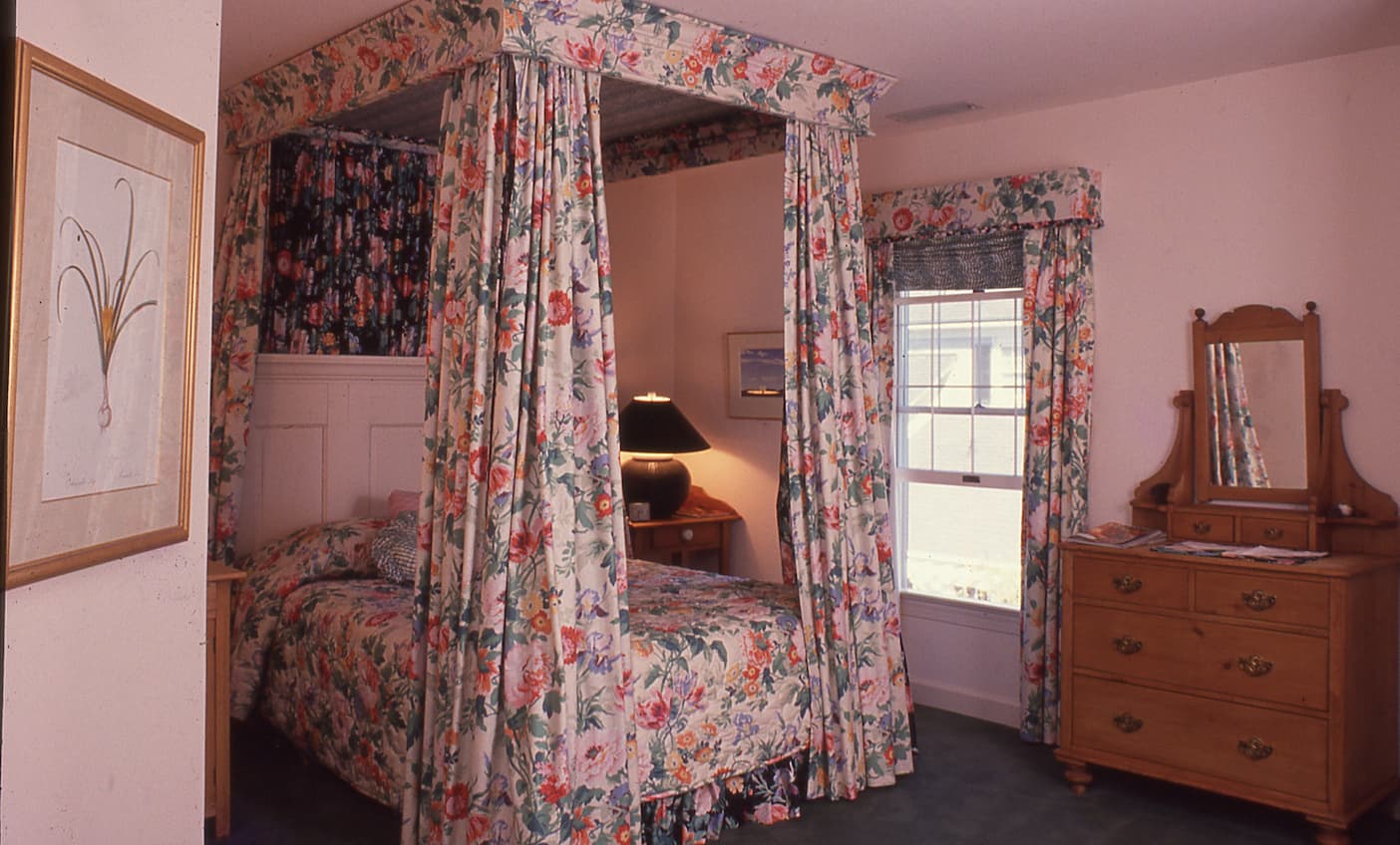 Guest room at the Fearrington Inn, decorated in 1988 by Jenny Fitch, featuring a floral canopy bed with matching draperies, a wooden dresser with mirror, framed artwork, and a bedside lamp beside a sunlit window.