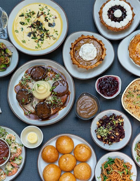 A table set with a variety of dishes including shrimp cocktail, bread rolls, mashed potatoes, meat with gravy, mixed vegetables, dessert pies, soups, and side sauces. Two empty wine glasses are placed on the table. The backdrop features greenery. Fearrington Village
