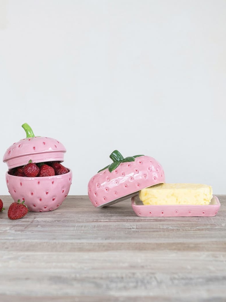 Two whimsical pink ceramic strawberry containers sit on a rustic wood tabletop against a soft gray background. One strawberry-shaped bowl is filled with fresh strawberries and topped with a matching lid, while the other serves as a butter dish, holding a stick of butter beneath a strawberry-shaped cover. The playful design combines vintage charm with functional kitchen storage.