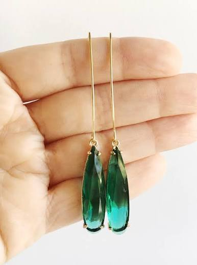 Long gold drop earrings featuring faceted emerald green teardrop stones, displayed against a hand. Fearrington Village.