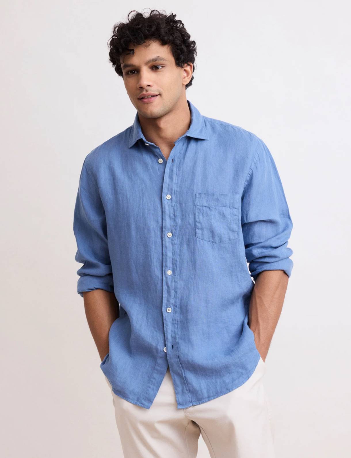 A man with short curly hair stands against a light background wearing a relaxed blue button-down shirt with sleeves rolled up and light-colored pants. His hands rest in his pockets, and he looks slightly to the side with a calm, casual expression.