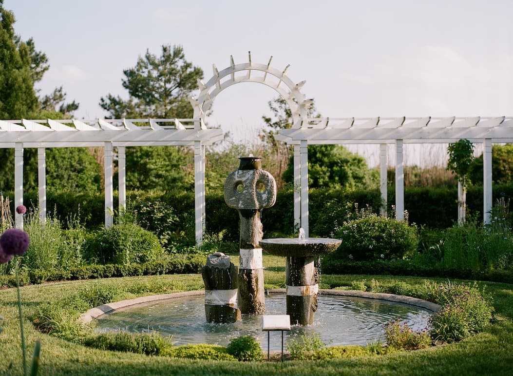 Formal garden with a circular fountain and sculptural stone centerpiece, framed by a white pergola and clipped hedges on a clear day.