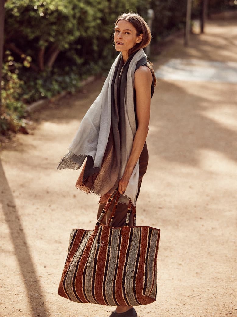 A woman walks along a sunlit garden path carrying a large woven tote bag with earthy striped patterns in rust, brown, gray, and cream tones. Draped in a soft ombré scarf and dressed in neutral layers, she showcases the bag’s generous size and artisanal texture. The relaxed, natural setting highlights the tote as a versatile accessory for travel, shopping, or everyday outings.