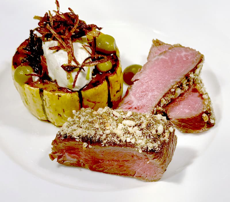 A gourmet dish featuring two cuts of seared rare beef, one topped with a crumb garnish, served with a stuffed, roasted vegetable ring filled with a creamy cheese, olives, and crispy fried garnish on a white plate. Fearrington Village