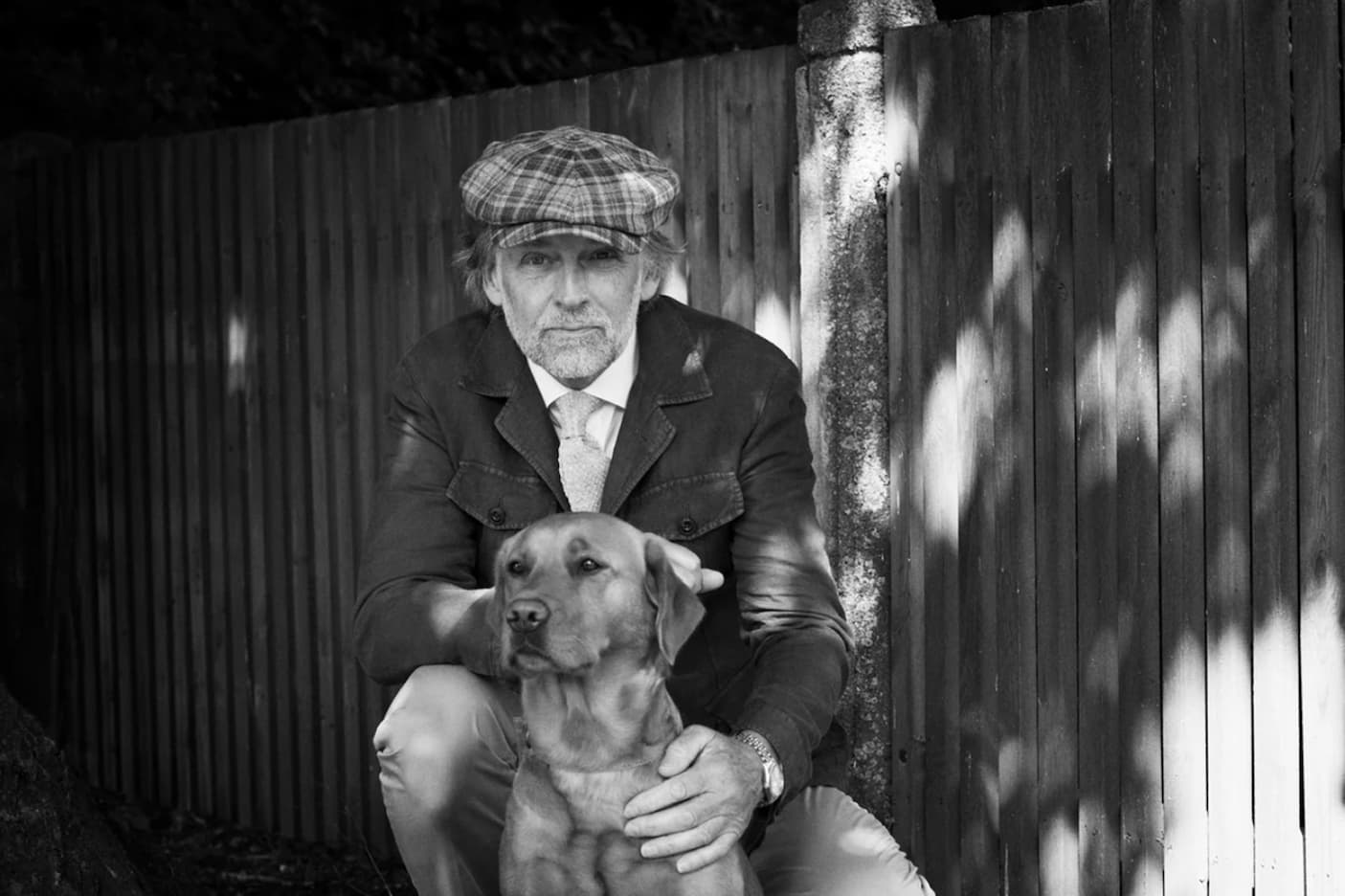 Black-and-white portrait of a man in a plaid cap posing outdoors with a Labrador retriever. Fearrington Village.