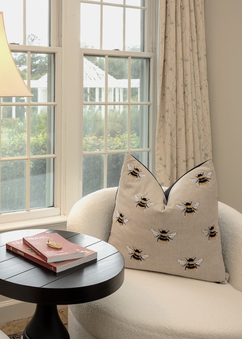 Cozy window-side seating area with a cream upholstered chair, a decorative pillow embroidered with bees, and a small round black table holding a couple of books; soft natural light filters through paned windows with neutral curtains.