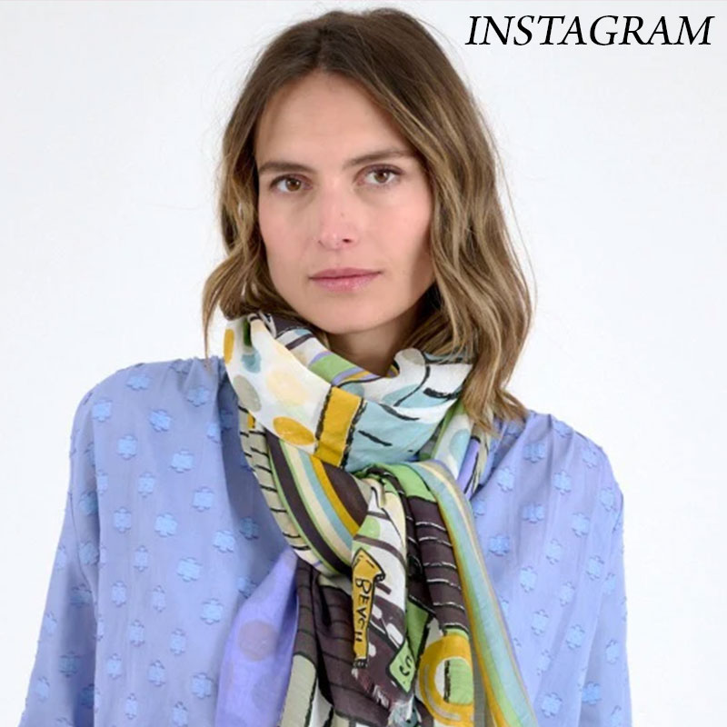 Woman wearing a light blue textured blouse styled with a multicolored patterned scarf wrapped around her neck, with “Instagram” text in the top corner against a neutral background.