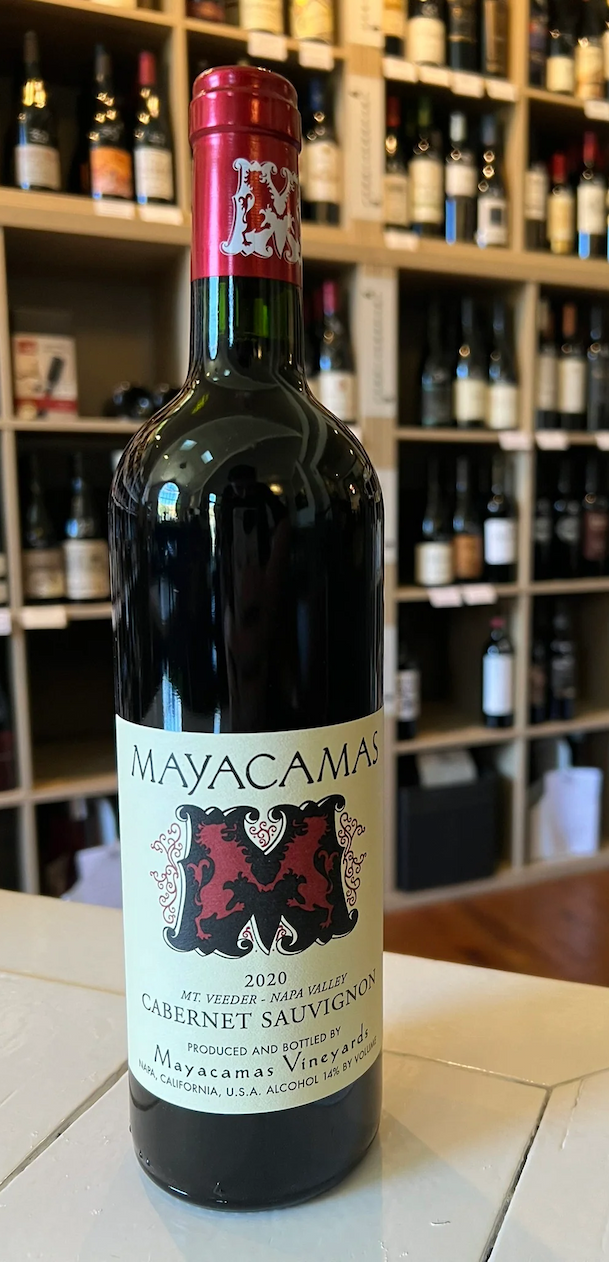 A bottle of 2020 Mayacamas Cabernet Sauvignon stands on a table in front of shelves filled with various wine bottles in a store. Fearrington Village