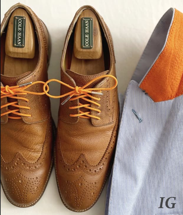 A pair of tan Cole Haan brogue dress shoes with bright orange laces beside a striped jacket with an orange collar accent.