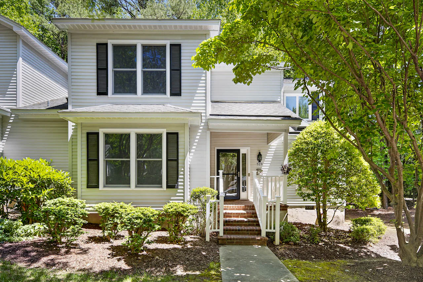 A bright two-story home with light siding and black shutters features a small front porch, brick steps, and a neatly landscaped yard with trimmed shrubs and trees in a sunny, residential setting.