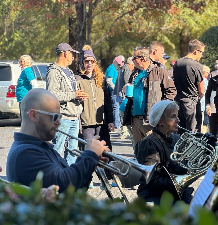 A group of people gather outdoors on a sunny day; some chat while others play musical instruments, including a trumpet and a French horn. Most are dressed in casual, warm clothing and sunglasses. Trees and parked cars are in the background. Fearrington Village