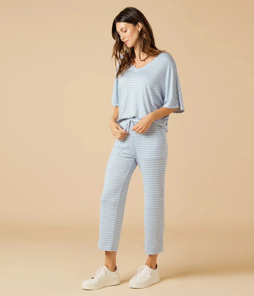 A model stands against a warm beige background wearing a soft blue-and-white striped lounge set featuring a relaxed V-neck top with draped sleeves and matching straight-leg drawstring pants, paired with white sneakers.