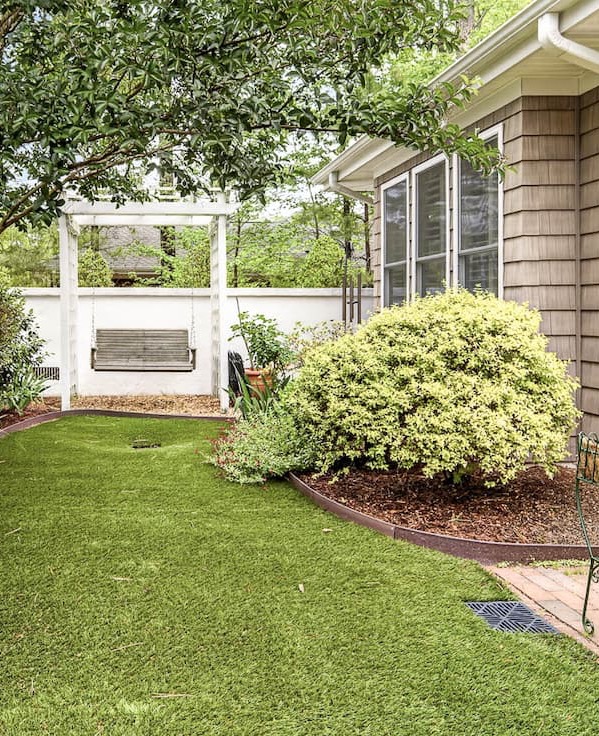 A beautifully landscaped backyard features a manicured putting green, mature trees, flowering shrubs, and a charming pergola with a porch swing, creating a peaceful outdoor retreat. Fearrington Village.