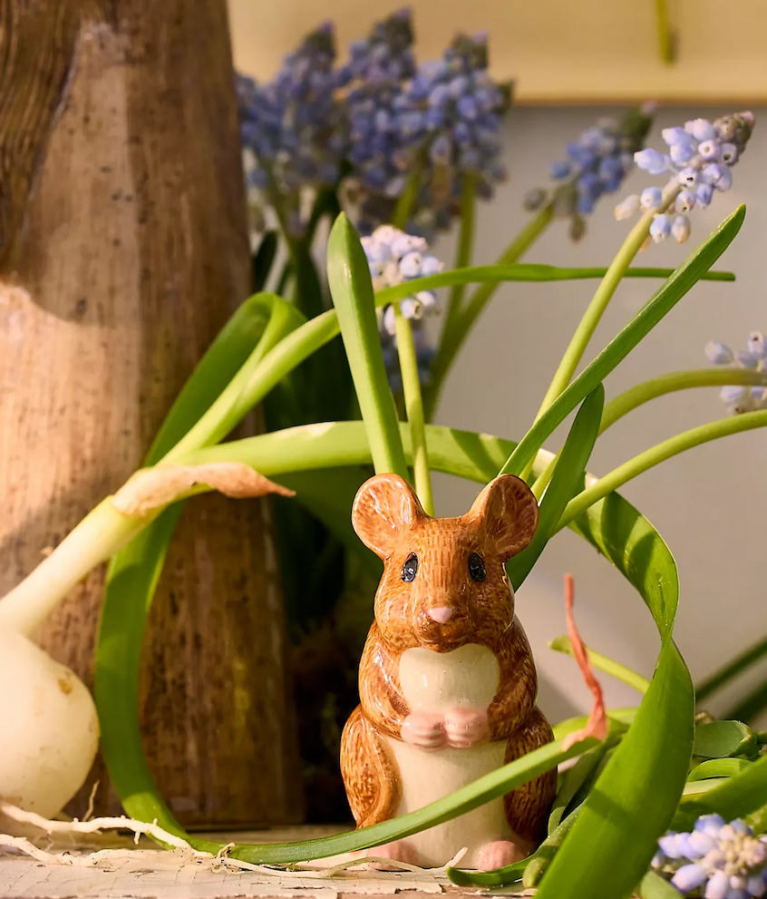 Small ceramic mouse figurine sits among green leaves and pale blue grape hyacinth flowers beside a wooden vase in warm sunlight.