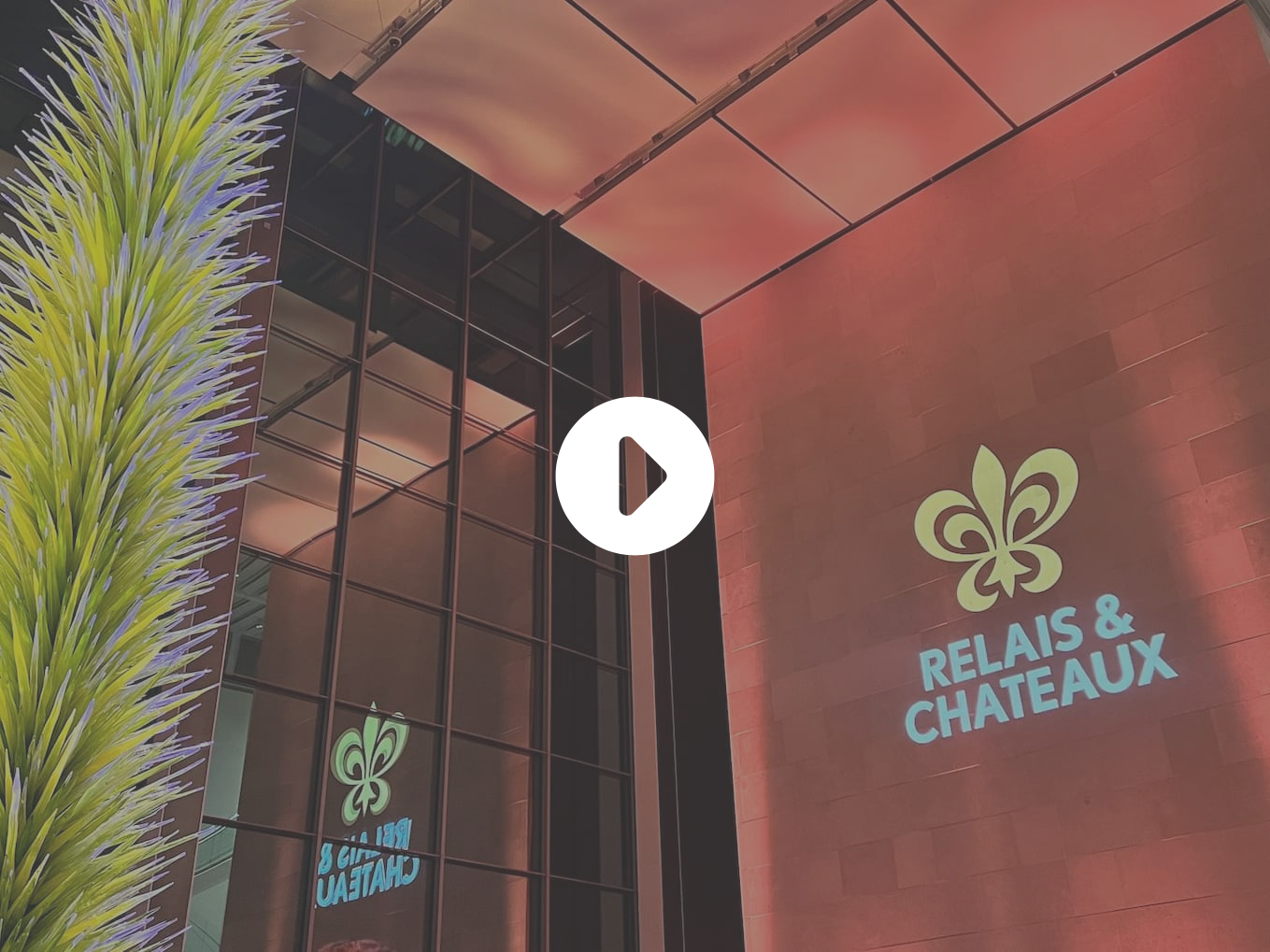 A modern building exterior with large glass panels, a tall decorative spiky green sculpture, and the “RELAIS & CHATEAUX” logo and emblem illuminated on a wall. Fearrington Village