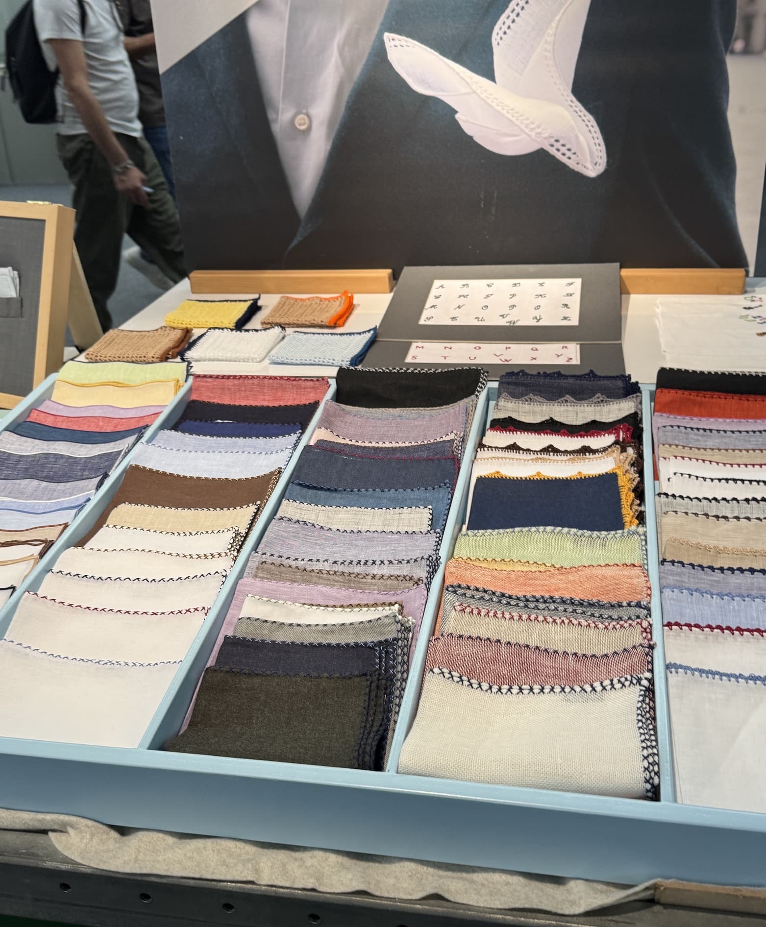 A tabletop display of neatly arranged fabric swatches and textile samples in soft neutrals, blues, pinks, and creams. The fabrics feature decorative stitched edges and sit beneath a large photograph of a white pocket square tucked into a dark suit jacket.