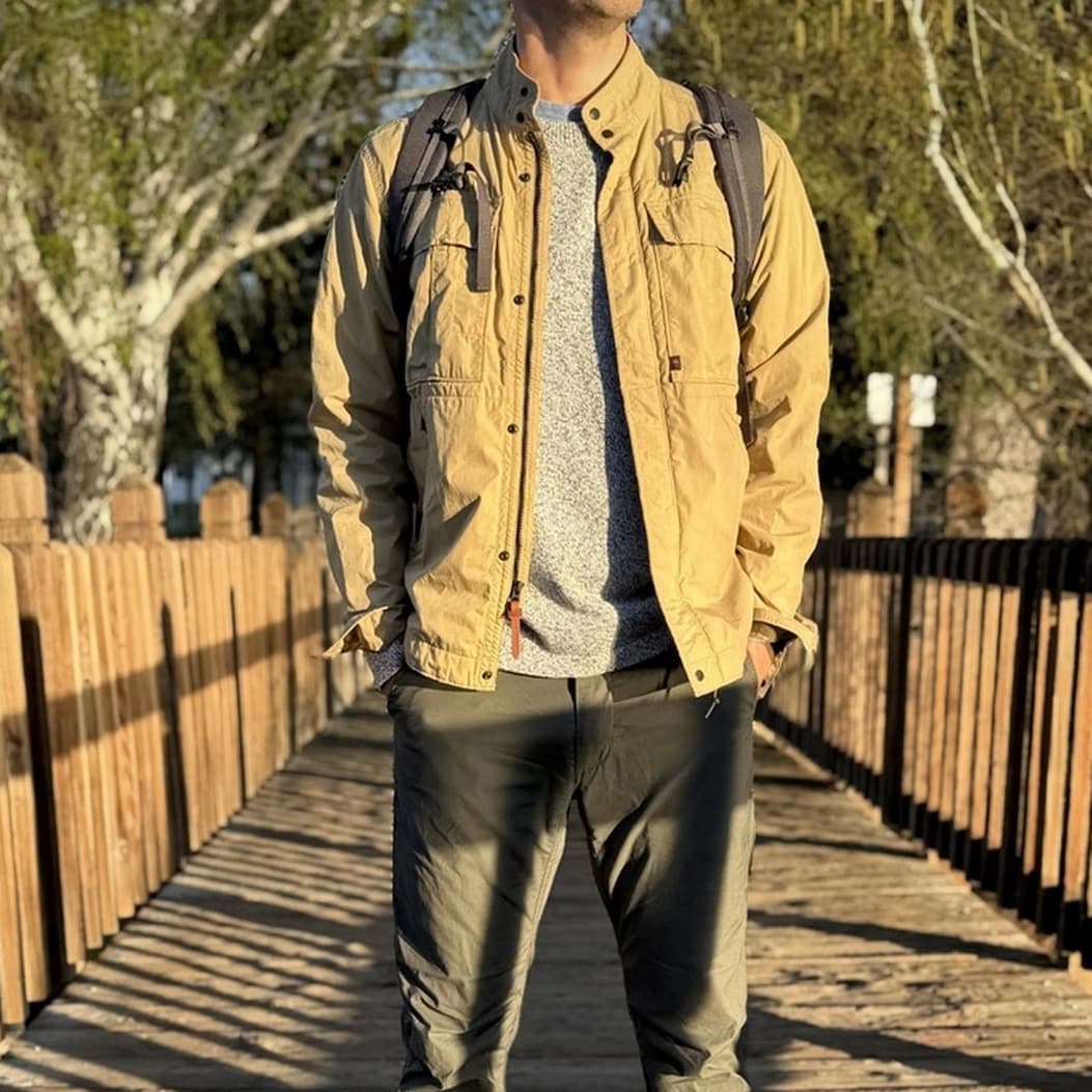 A casually dressed man stands on a sunlit wooden walkway wearing a light tan field jacket layered over a heather gray sweater, dark relaxed-fit trousers, and a backpack. Warm golden-hour light highlights the textures of the clothing, while the tree-lined path and long shadows create an outdoor, adventure-ready feel. The outfit combines practical comfort with understated style, making it well suited for travel, weekend walks, or everyday exploration.