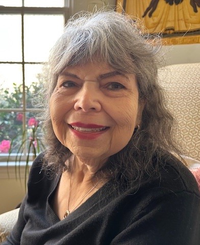 An older woman with gray hair smiles while seated indoors near a window, with soft natural light and flowers visible outside.